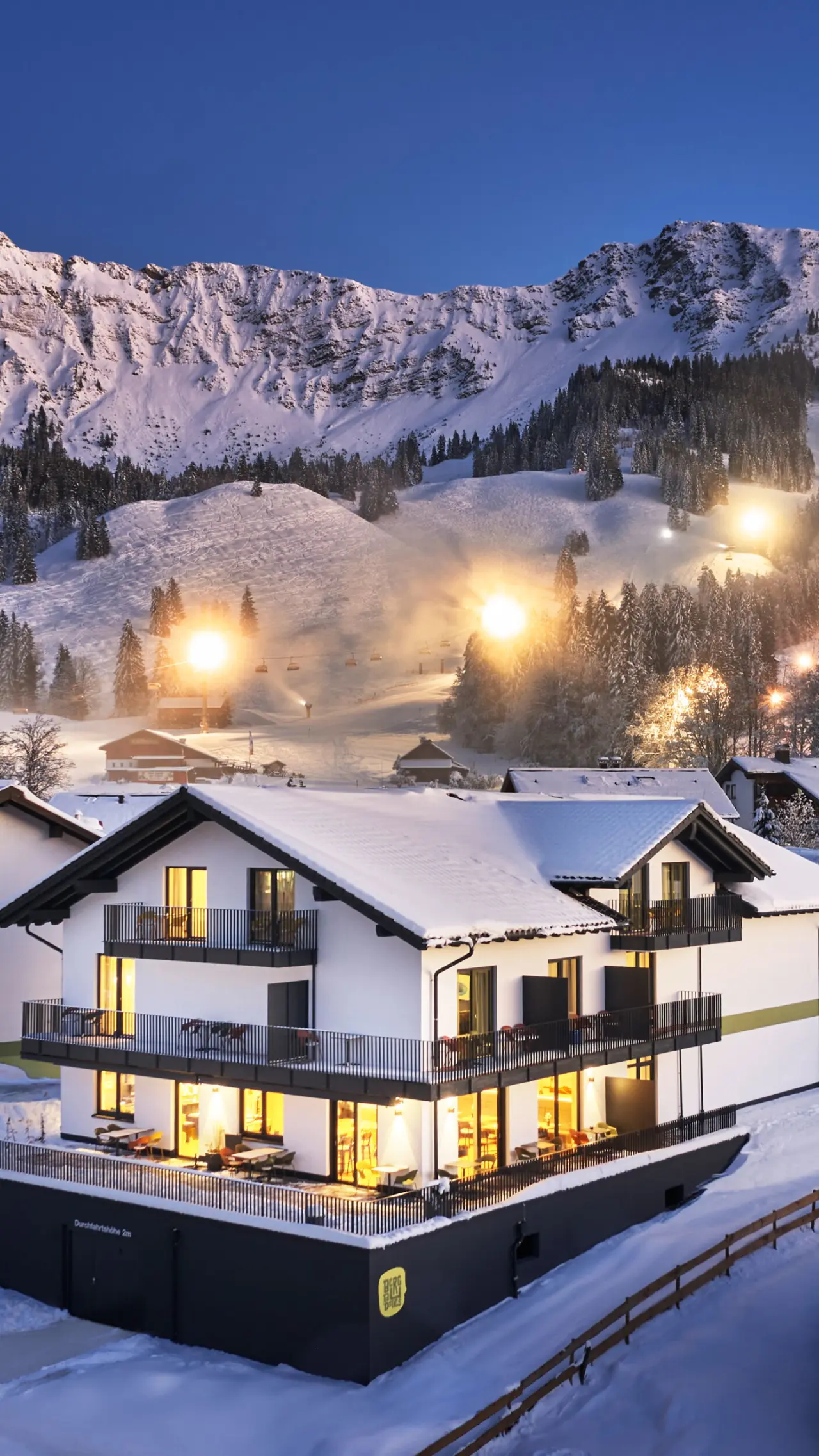 Modernes Hotel BergBuddies bei Dämmerung im Schnee mit beleuchteten Balkonen und verschneiten Bergen im Hintergrund.
