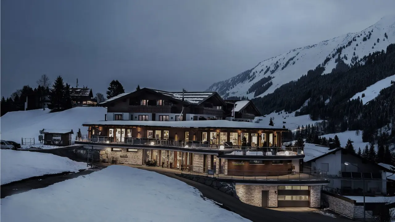 Beleuchtetes Hotel Birkenhöhe in verschneiter Alpenlandschaft bei Nacht, umgeben von Bergen und Tannen.