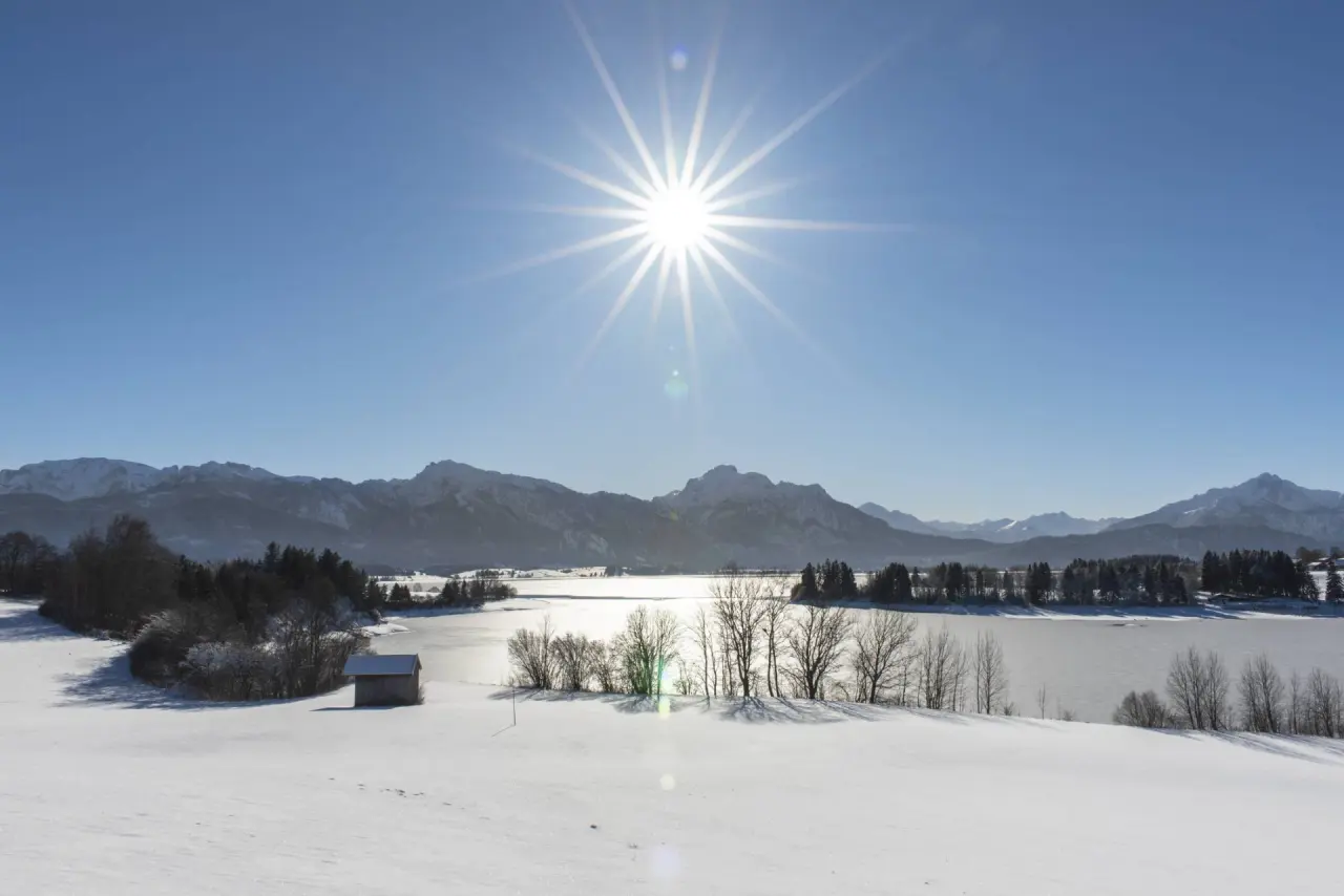 Winterlandschaft mit verschneiten Feldern, zugefrorenem See und Alpenpanorama unter strahlendem Sonnenschein bei Hartung’s Hoteldorf.