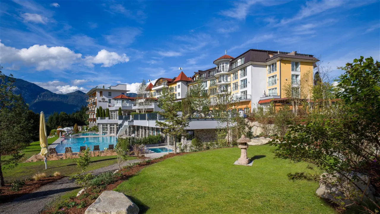 Aussenansicht des Hotel Panorama Royal mit grossem Aussenpool, Liegewiese und Bergpanorama unter blauem Himmel.