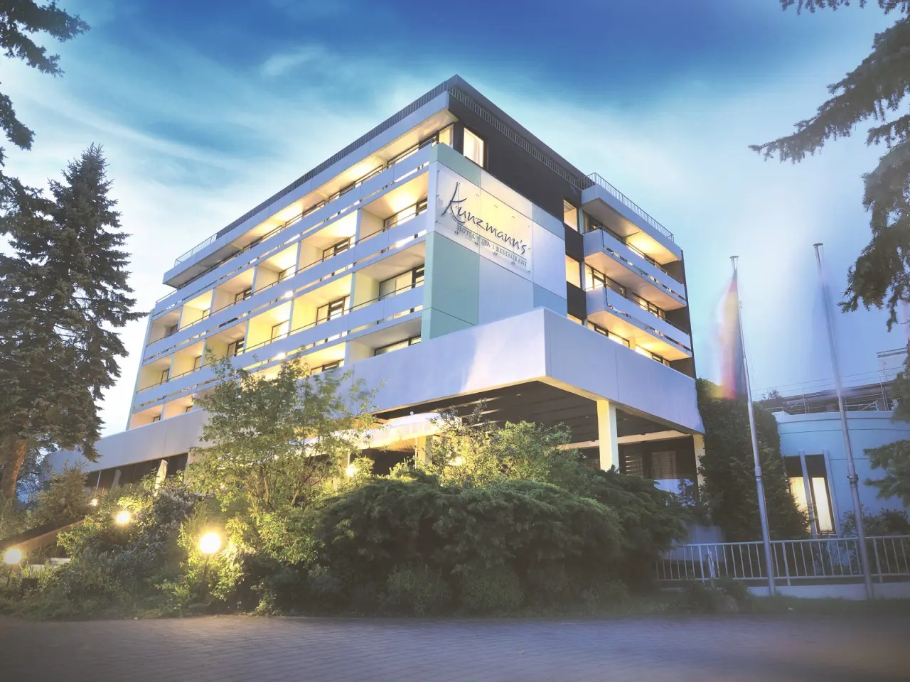 Abendansicht des Kunzmann's Hotel | Spa mit beleuchteter Fassade und Balkonen unter blauem Himmel.