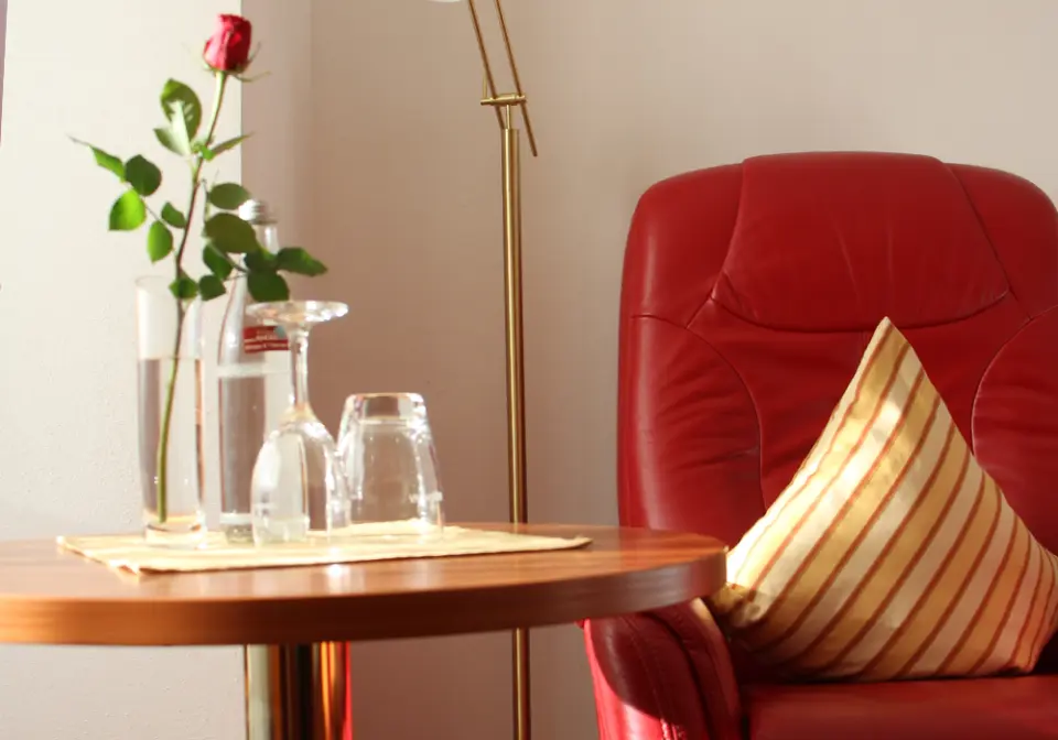 Gemütliche Sitzecke im ANGERHOF Kur- und Sporthotel mit rotem Sessel, Beistelltisch und frischer Rose.
