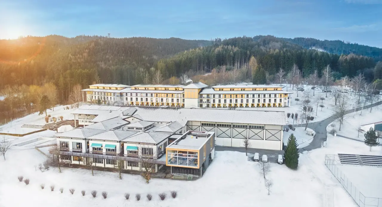 Luftaufnahme des Aldiana Club Ampflwang im Winter, umgeben von verschneiten Wäldern und Hügeln unter blauem Himmel.