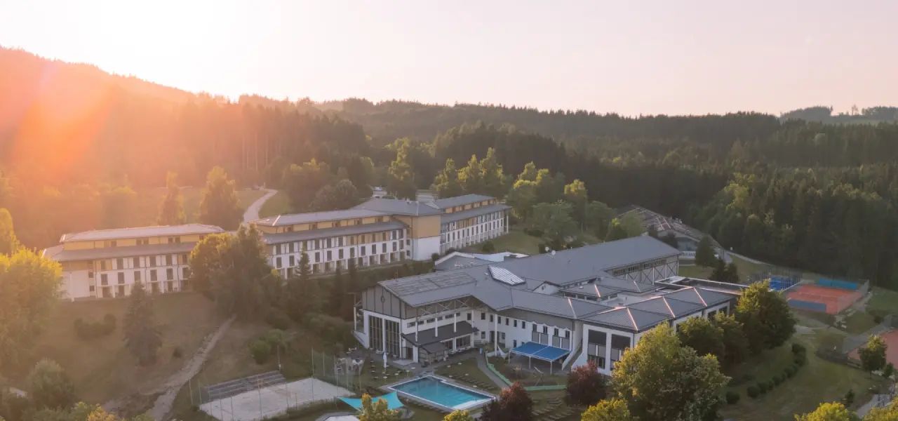 Luftaufnahme des Aldiana Club Ampflwang mit Hotelgebäuden, Aussenpool und Sportplätzen inmitten grüner Wälder bei Sonnenuntergang.