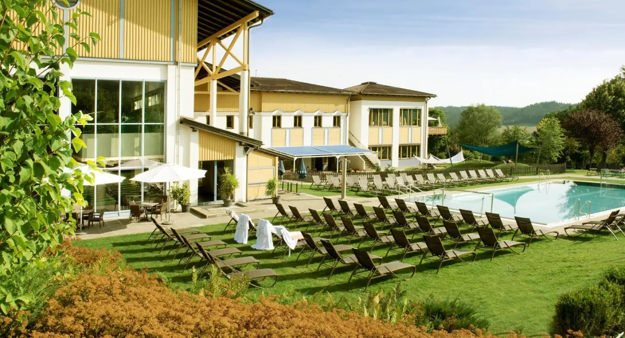 Aussenansicht des Aldiana Club Ampflwang mit grossem Aussenpool, Liegewiese und Hotelgebaeude unter blauem Himmel.