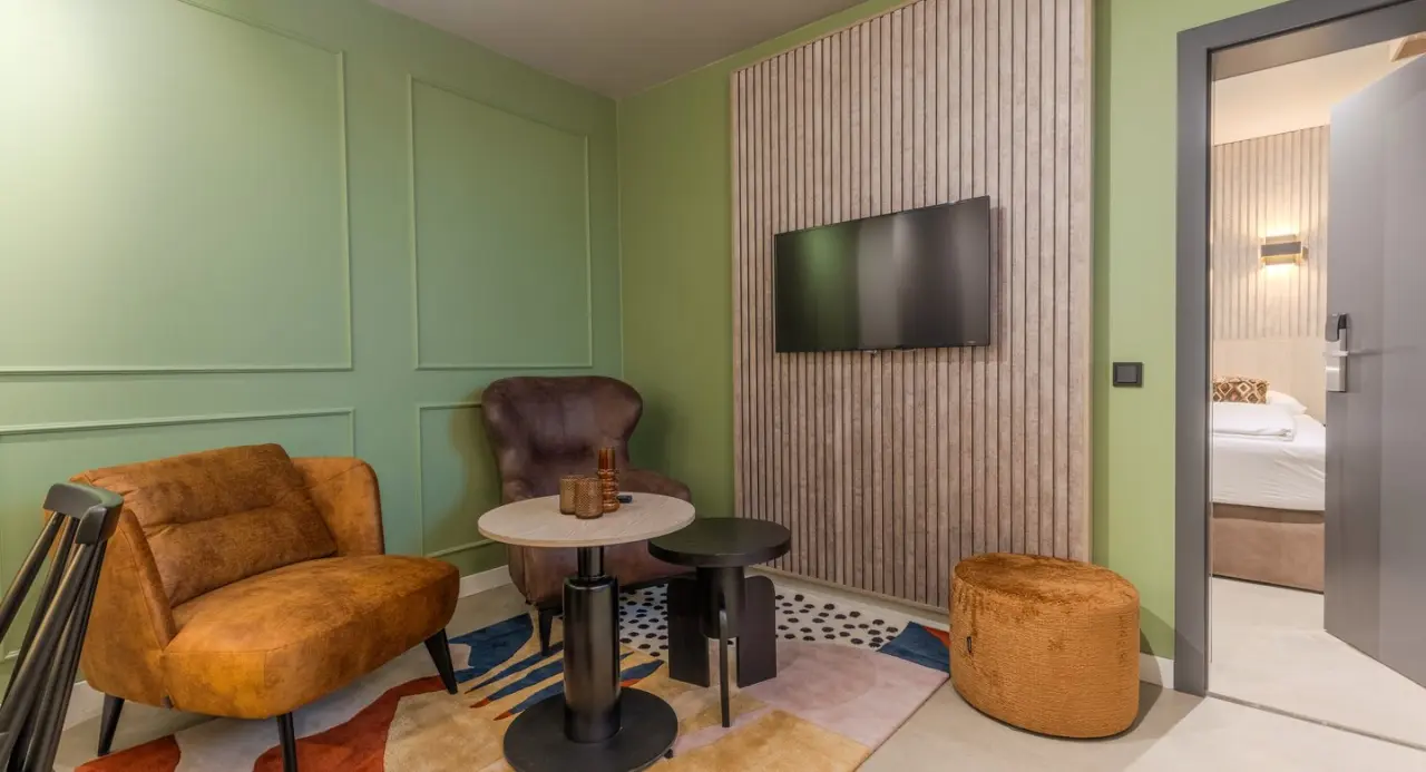 Gemütlicher Wohnbereich in einem Hotelzimmer des Aldiana Club Ampflwang mit Sessel, Couch, TV und Blick ins Schlafzimmer.