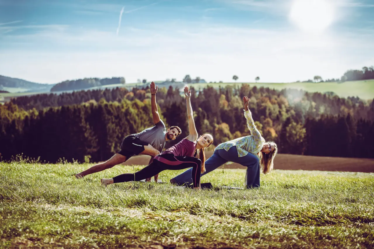 Drei Personen praktizieren Yoga in der Natur mit Blick auf die Hügellandschaft des Landrefugium Obermüller.
