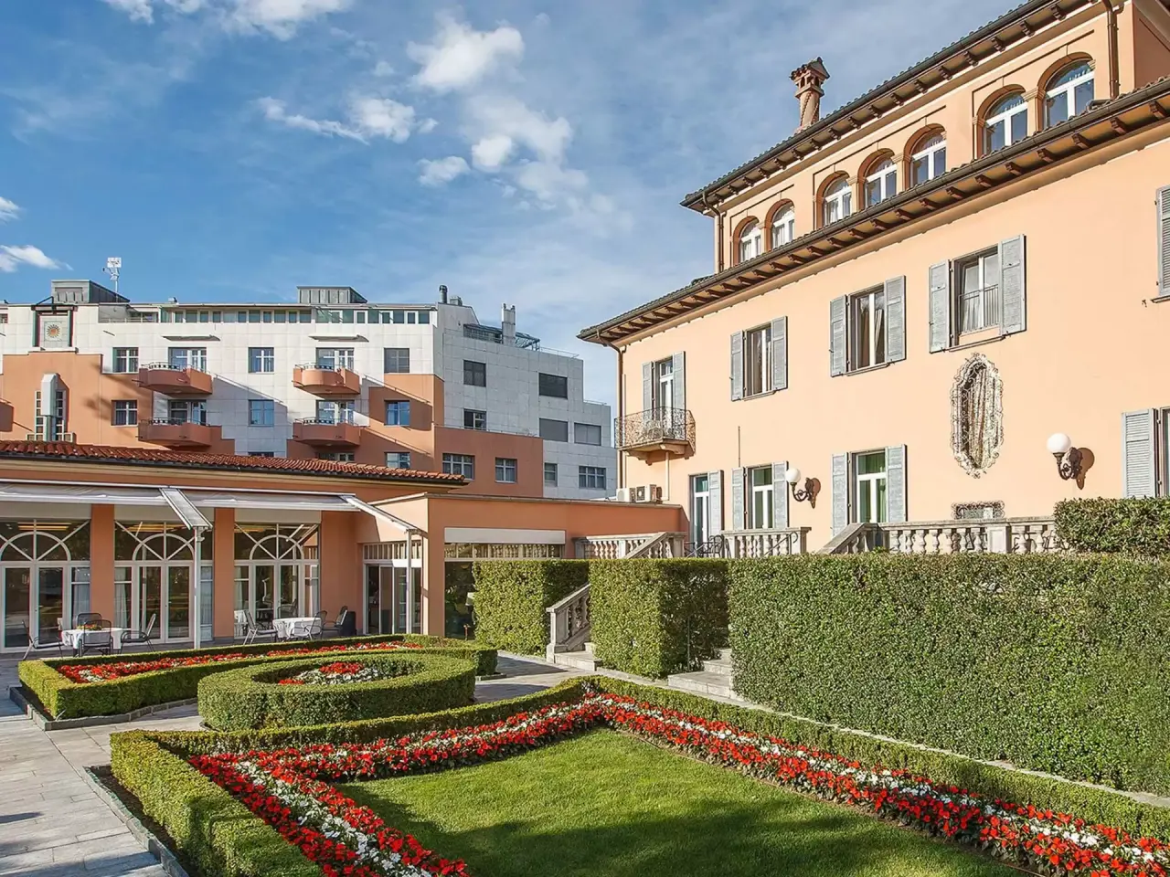 Aussenansicht des Villa Sassa Hotel Residence & SPA mit gepflegtem Garten und blühenden Blumenbeeten unter blauem Himmel.