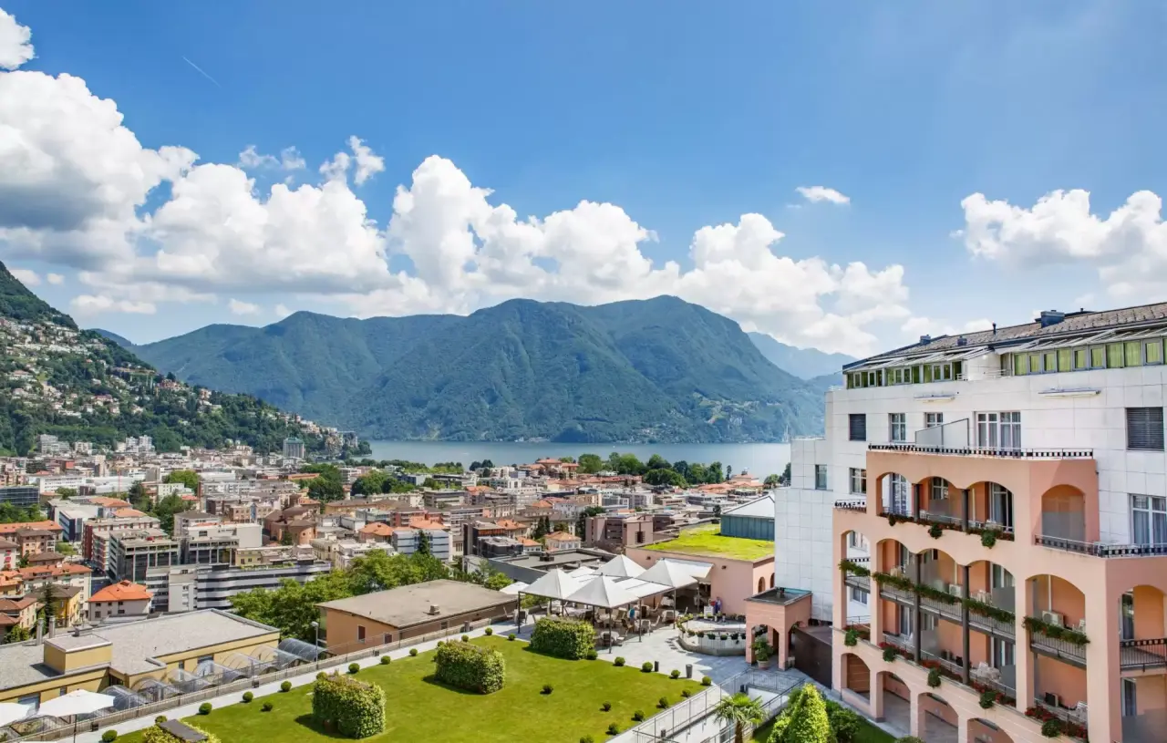 Panoramablick auf das Villa Sassa Hotel Residence & SPA mit Luganersee, Stadt und Bergen unter blauem Himmel.