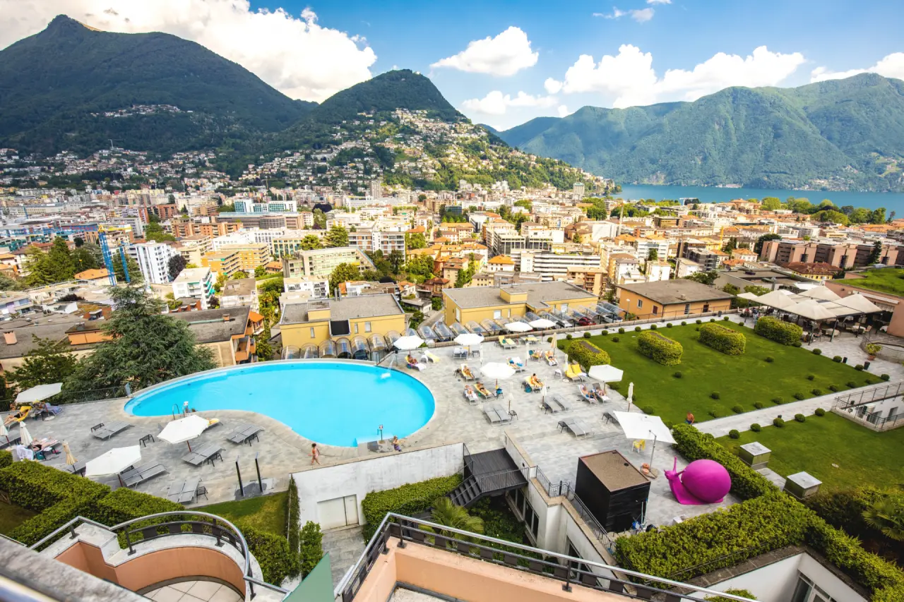 Panoramablick auf den Aussenpool der Villa Sassa Hotel Residence & SPA mit Luganersee und Bergen im Hintergrund.