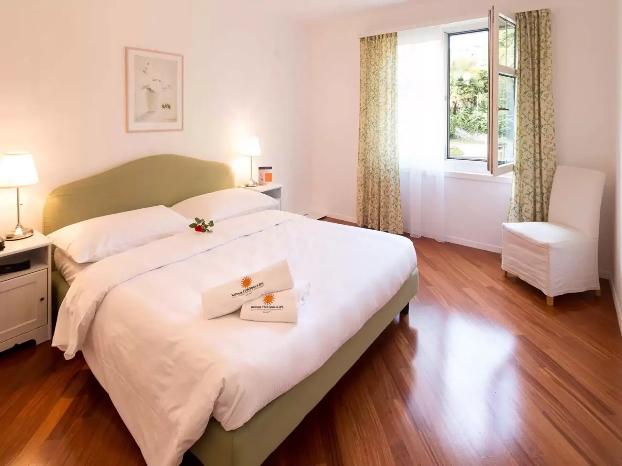 Helles Doppelzimmer mit Holzboden und geöffnetem Fenster im Villa Sassa Hotel Residence & SPA. Komfortables Bett mit Wellness-Handtüchern.