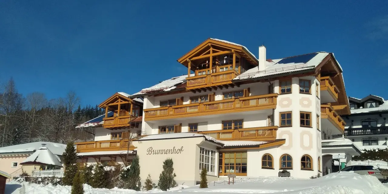 Winterliche Aussenansicht des Hotel Brunnenhof mit verschneiten Dächern und Holzbalkonen unter strahlend blauem Himmel.