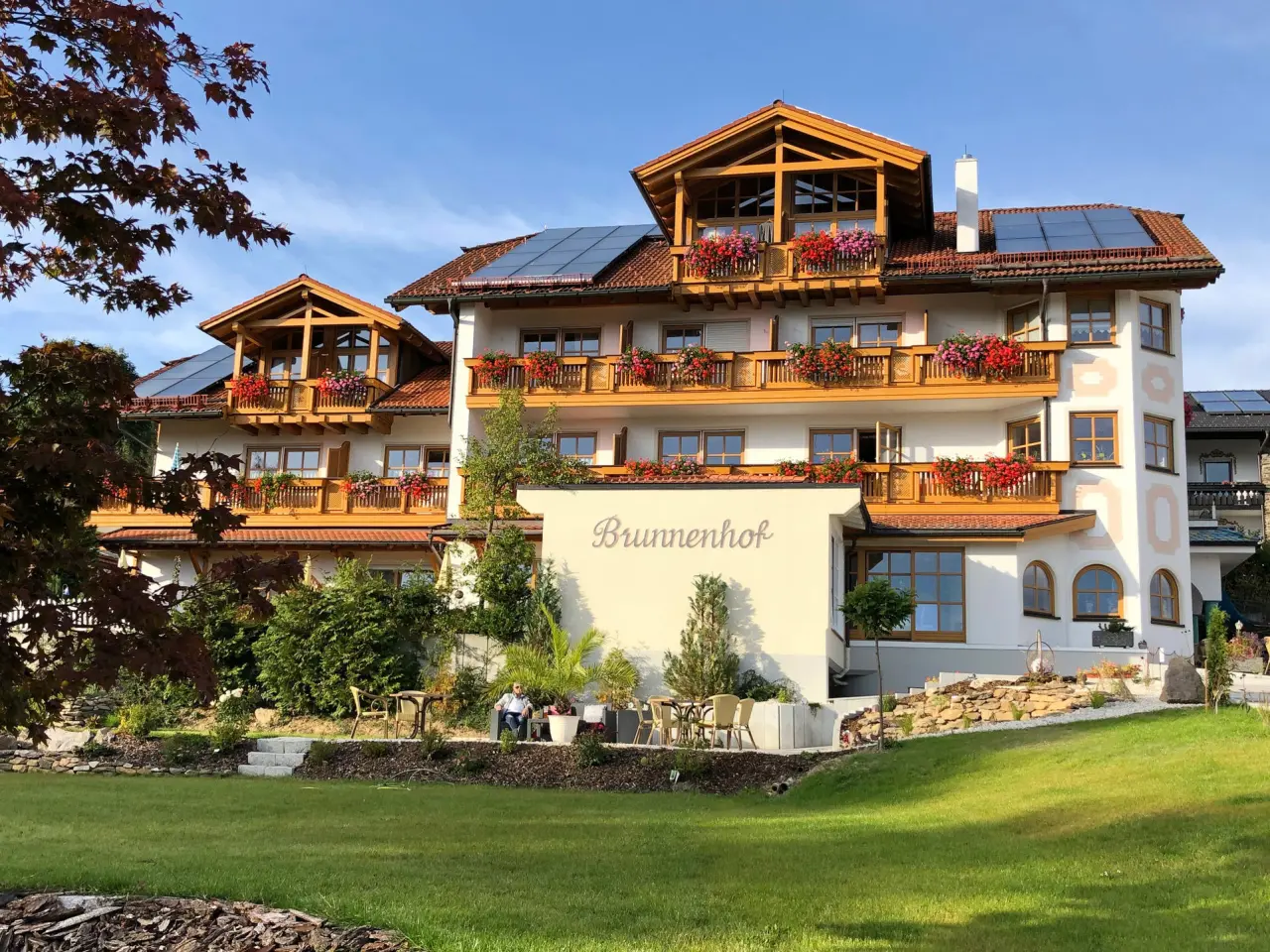Aussenansicht des Hotel Brunnenhof mit traditioneller Architektur, blumengeschmückten Balkonen und gepflegtem Garten unter blauem Himmel.