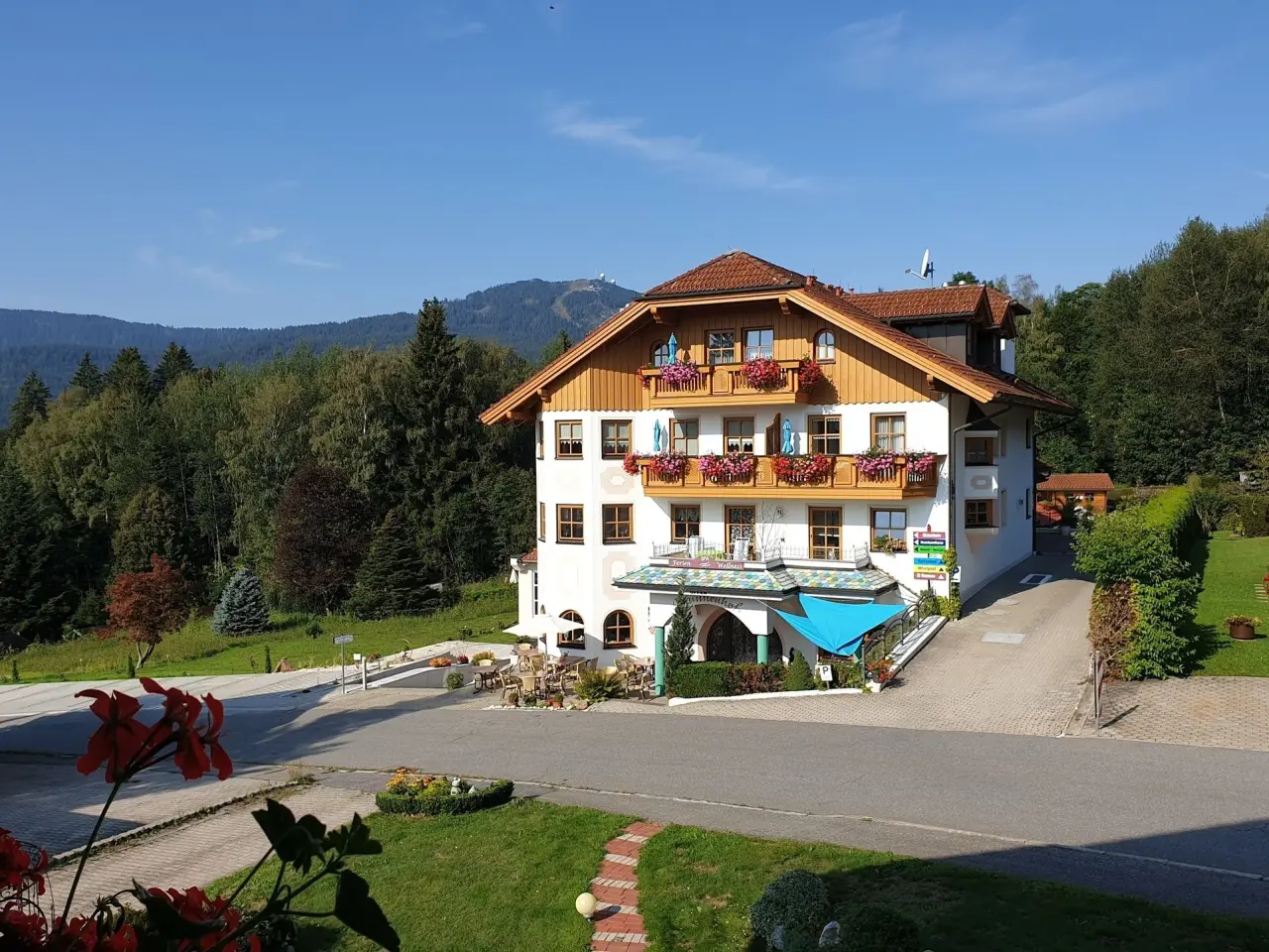 Aussenansicht des Hotel Brunnenhof in den Bergen, umgeben von Wald, mit blühenden Balkonen unter blauem Himmel.