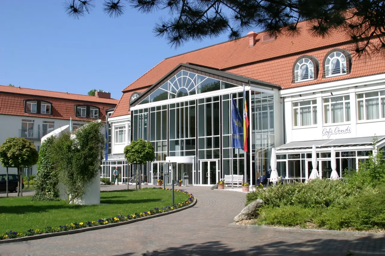 Aussenansicht des Seehotel Grossherzog v. Mecklenburg mit Glasfassade, rotem Dach und gepflegtem Garten bei Sonnenschein.