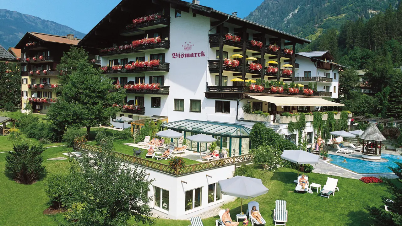Aussenansicht des Verwöhnhotel Bismarck mit blühenden Balkonen, großem Garten, Außenpool und Liegewiese vor Bergkulisse.
