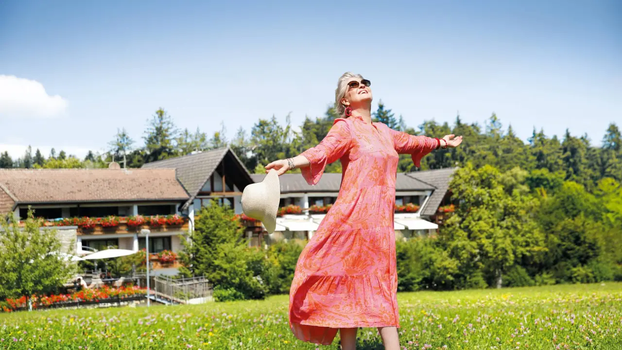 Lachende Frau genießt die Sonne auf einer blühenden Wiese vor dem Hotel Grüner Wald. Entspannung in der Natur.