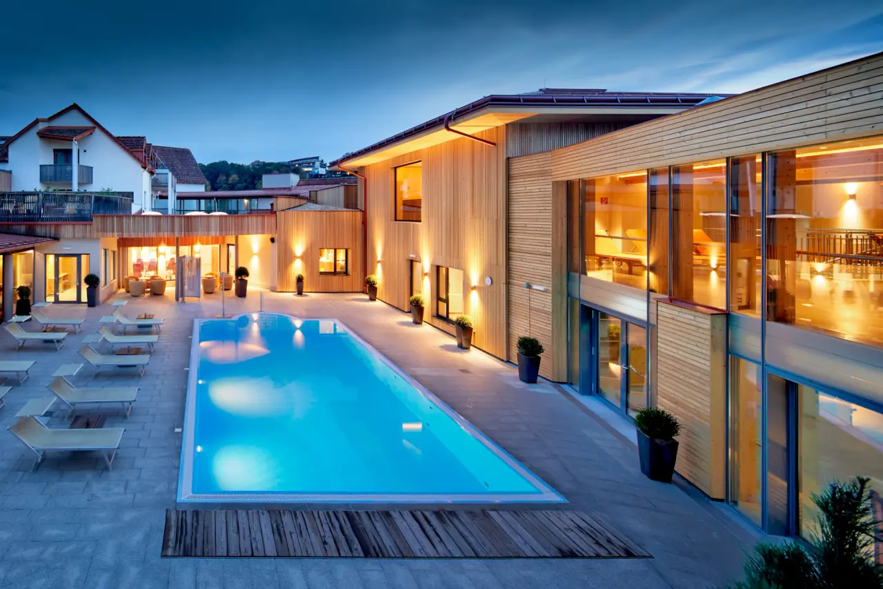Abendliche Aussenansicht des Ayurveda Resort Mandira mit beleuchtetem Pool und moderner Holzarchitektur.