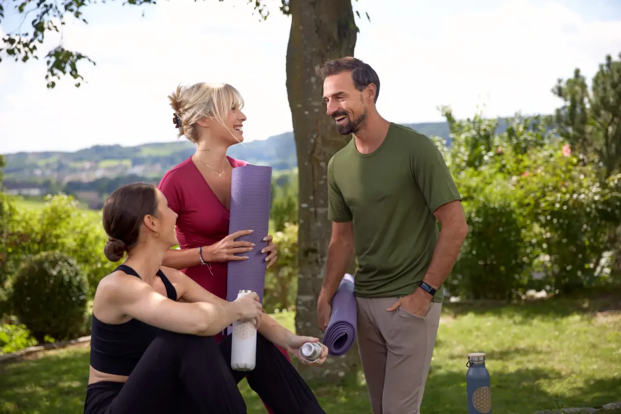 Drei lachende Gäste mit Yogamatten und Trinkflaschen im grünen Garten des Ayurveda Resort Mandira, bereit für eine Yoga-Session.