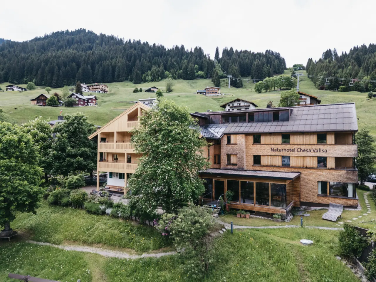 Naturhotel Chesa Valisa in den österreichischen Alpen, umgeben von grünen Wiesen und Wäldern, mit Bergbahn im Hintergrund.
