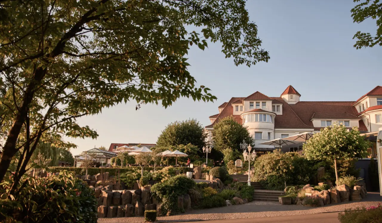 Aussenansicht des charmanten Hotel Heinz mit rotem Dach, umgeben von einem gepflegten Garten und sonniger Terrasse.