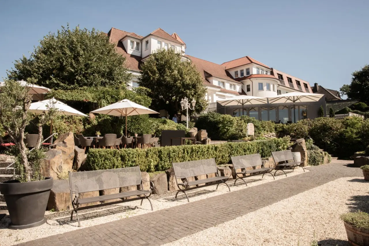 Aussenansicht des Hotel Heinz mit sonniger Gartenterrasse, Schirmen und Sitzgelegenheiten unter blauem Himmel.