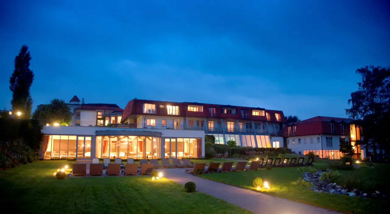 Abendliche Aussenansicht des Hotel Heinz mit beleuchtetem Gebäude, Garten und Liegestühlen unter blauem Himmel.