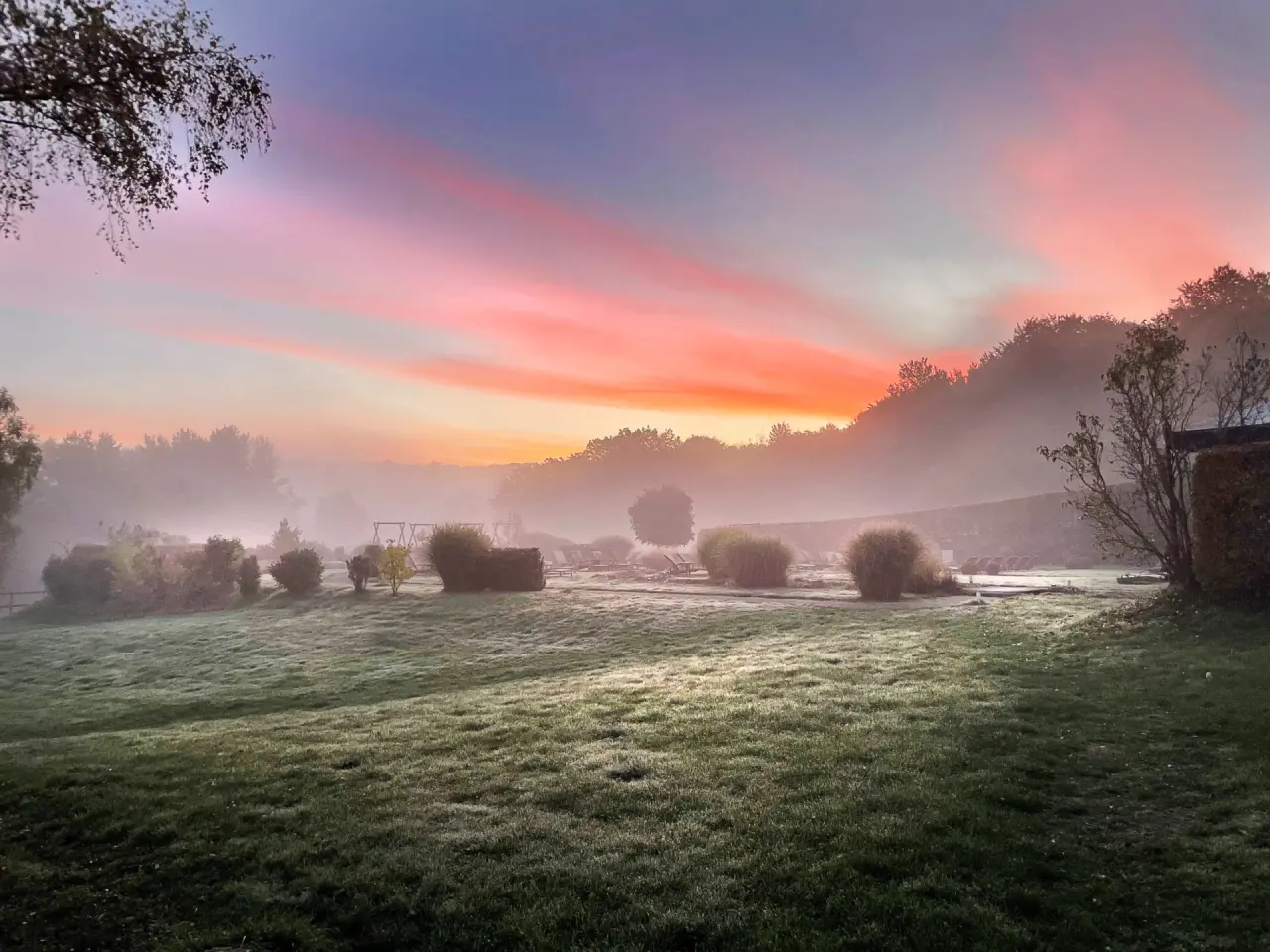 Malerischer Hotelgarten des Hotel Heinz im Morgennebel mit farbenprächtigem Sonnenaufgang und Tautropfen auf dem Gras.