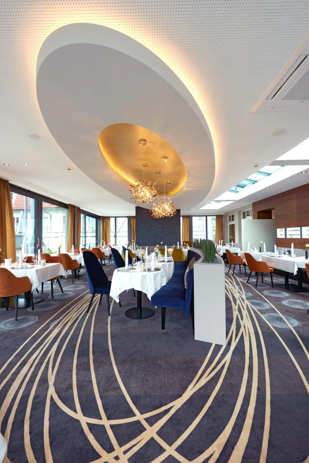 Modernes Restaurant mit stilvoller Einrichtung, blauen und orangen Stühlen, eleganten Tischen und goldener Deckenbeleuchtung im Hotel Heinz.