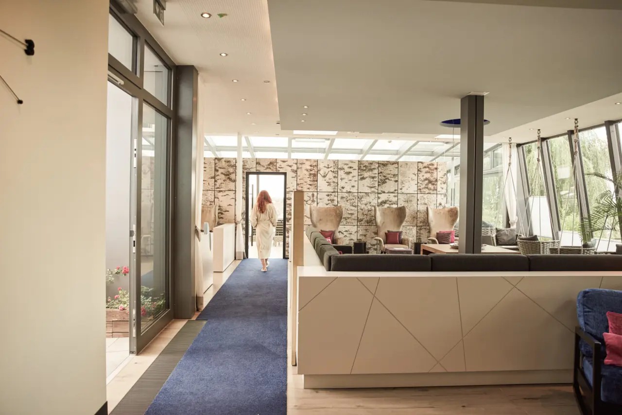 Heller Entspannungsbereich im Hotel Heinz mit Gast im Bademantel, gemütlichen Sesseln und Tageslichtdach.