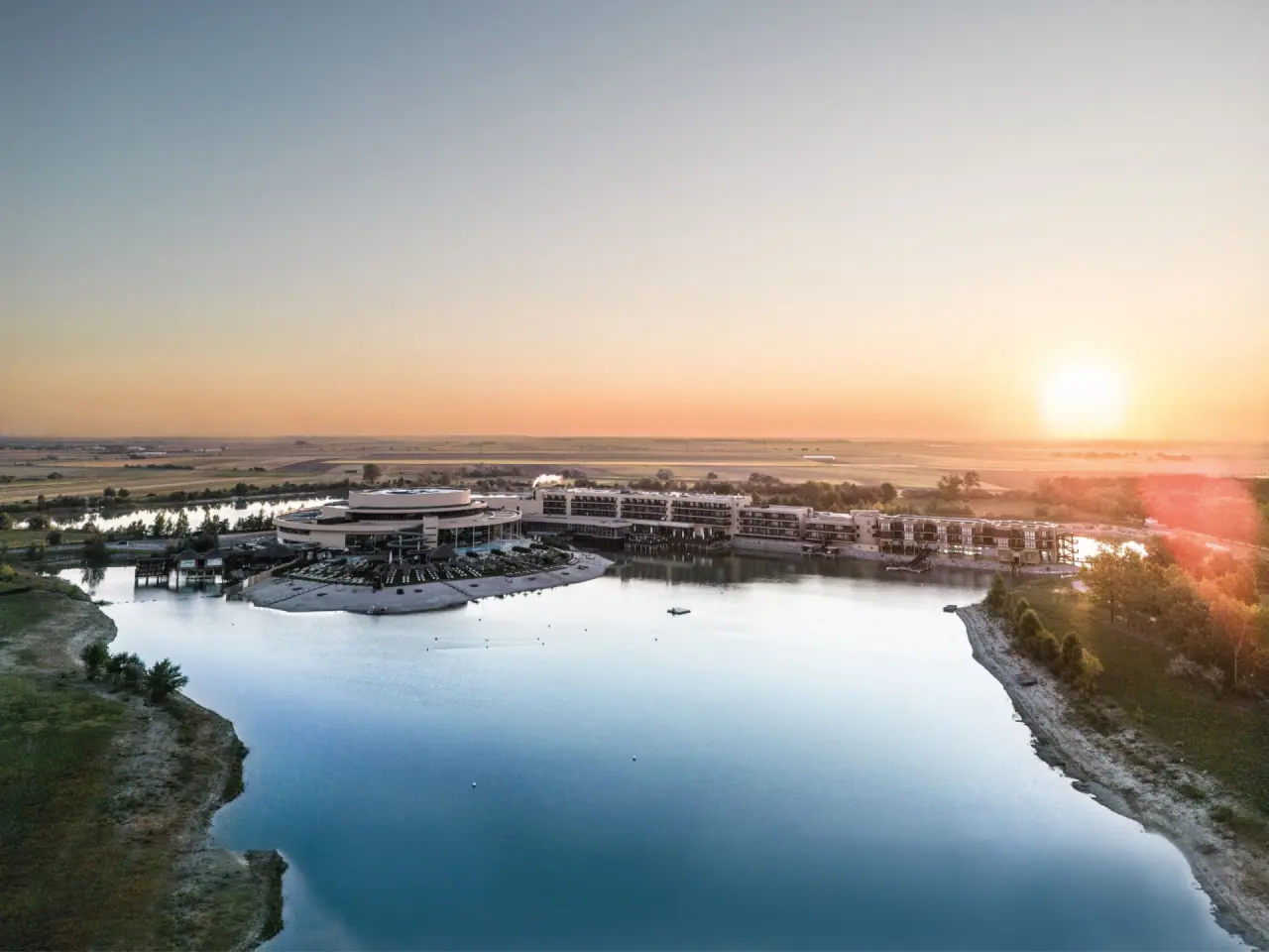 Panoramablick auf die St. Martins Therme & Lodge bei Sonnenuntergang, mit Hotelanlage, See und umliegender Natur.