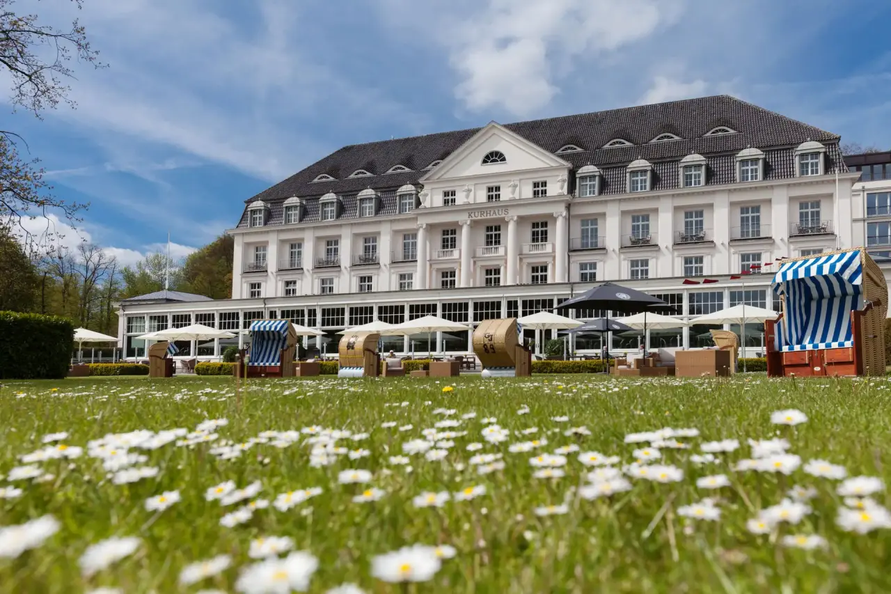 Grandhotel A-Rosa Kurhaus Travemünde mit blühender Wiese und Strandkörben unter blauem Himmel