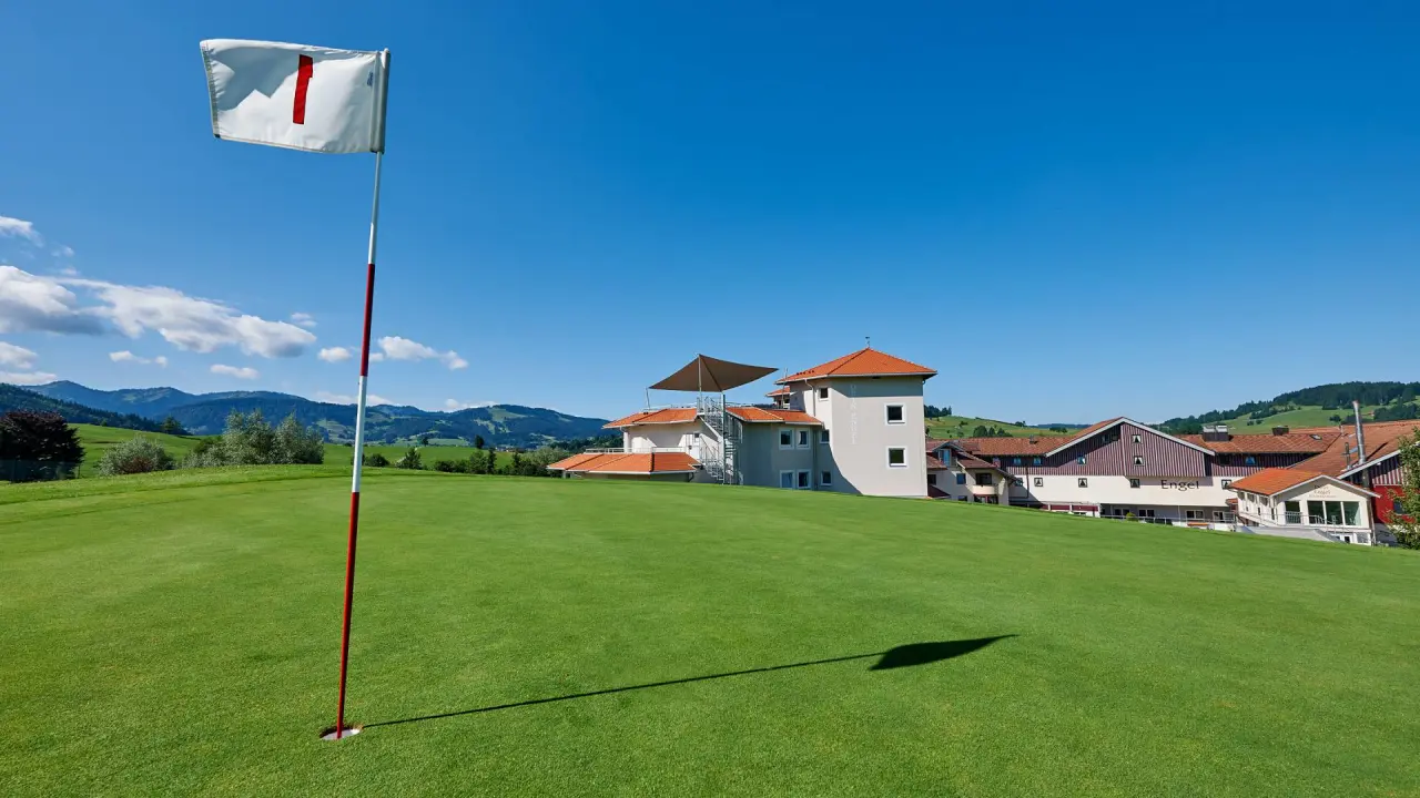 Golfplatz mit Fahne und Hotel Dein Engel im Hintergrund, umgeben von grünen Hügeln unter blauem Himmel.
