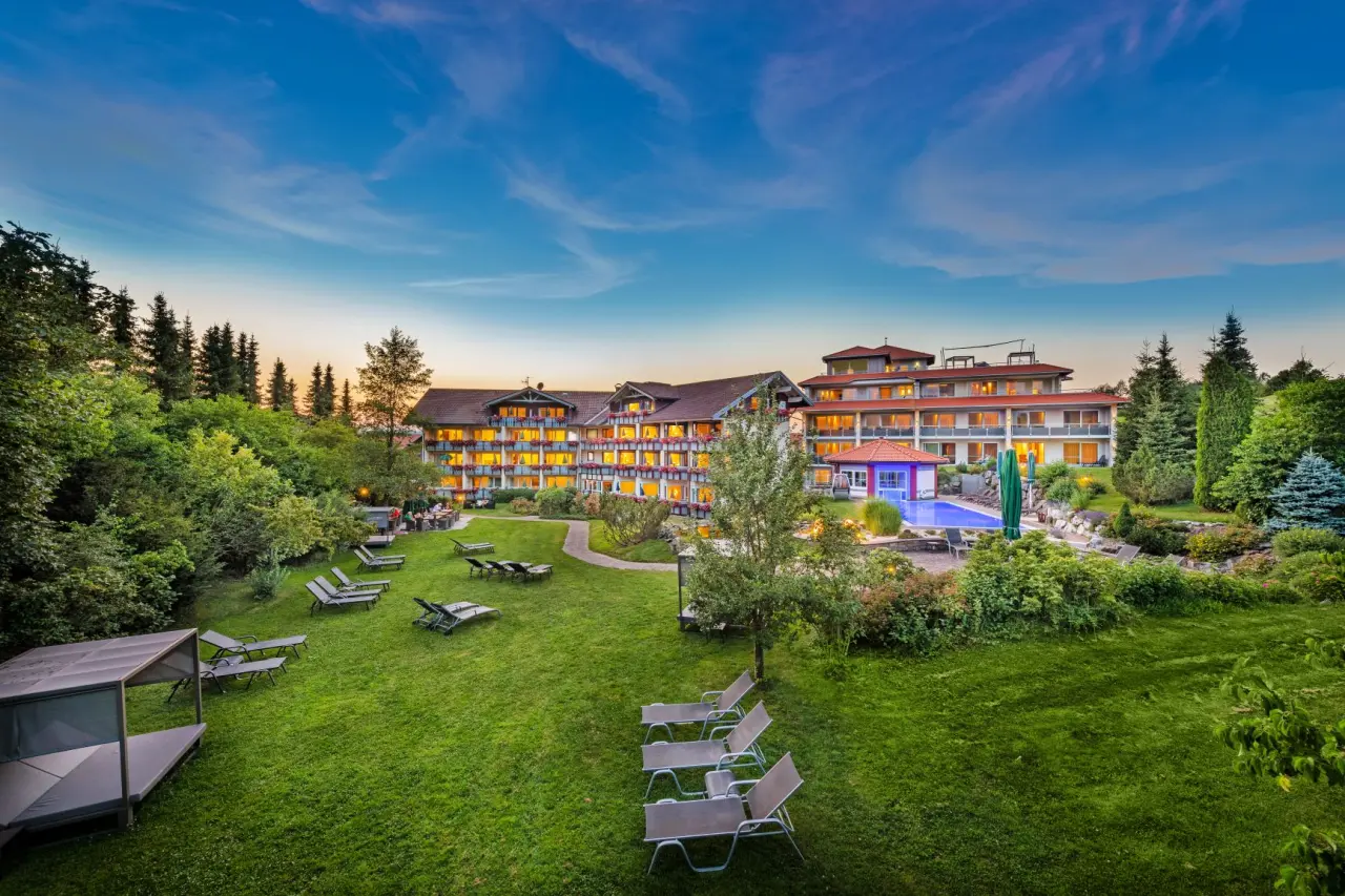 Abendansicht des Wellnesshotels Dein Engel mit beleuchteter Fassade, grossem Garten und Aussenpool unter blauem Himmel.