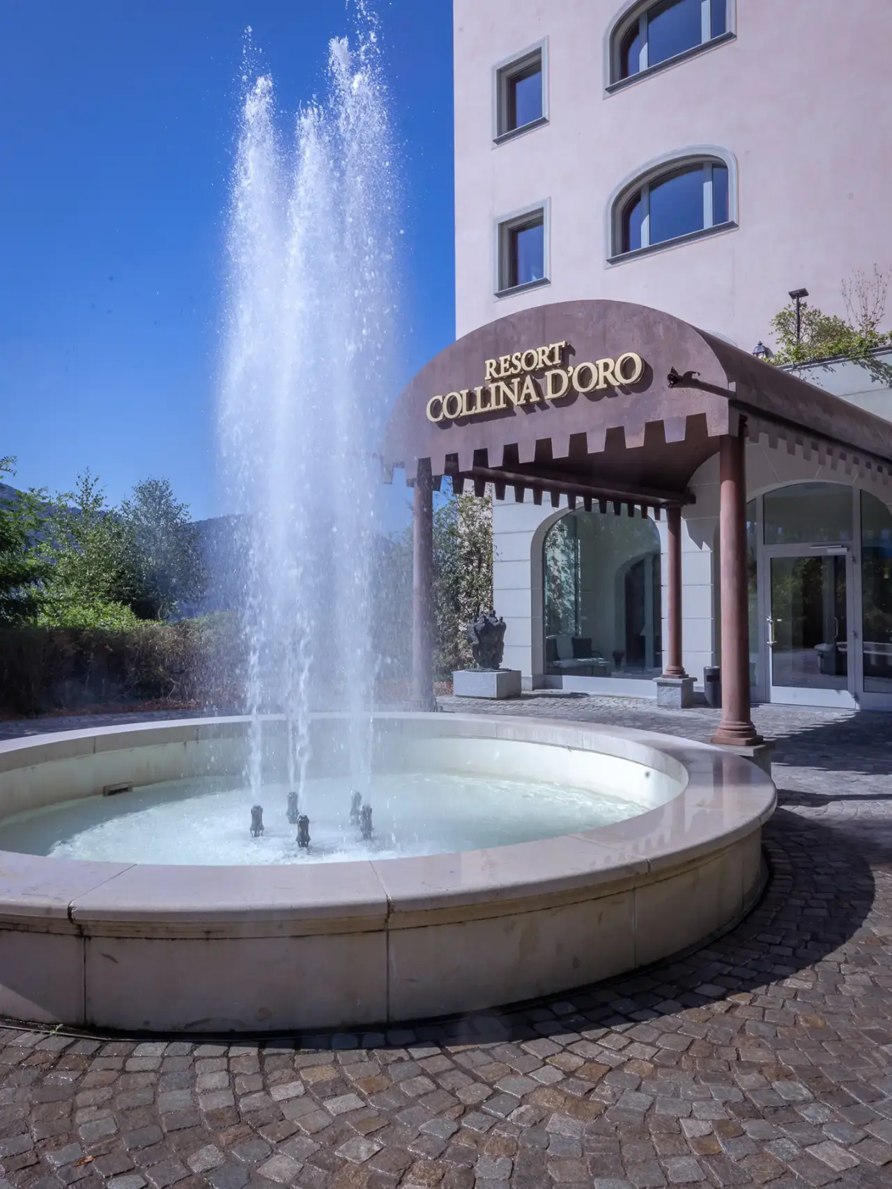 Eingangsbereich des Resort Collina d'Oro mit elegantem Springbrunnen und dem Hotelnamen an der Fassade.