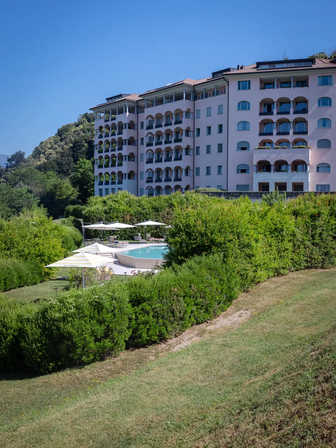 Aussenansicht des Resort Collina d'Oro mit rosa Fassade, umgeben von grünen Gärten und einem Aussenpool unter blauem Himmel.