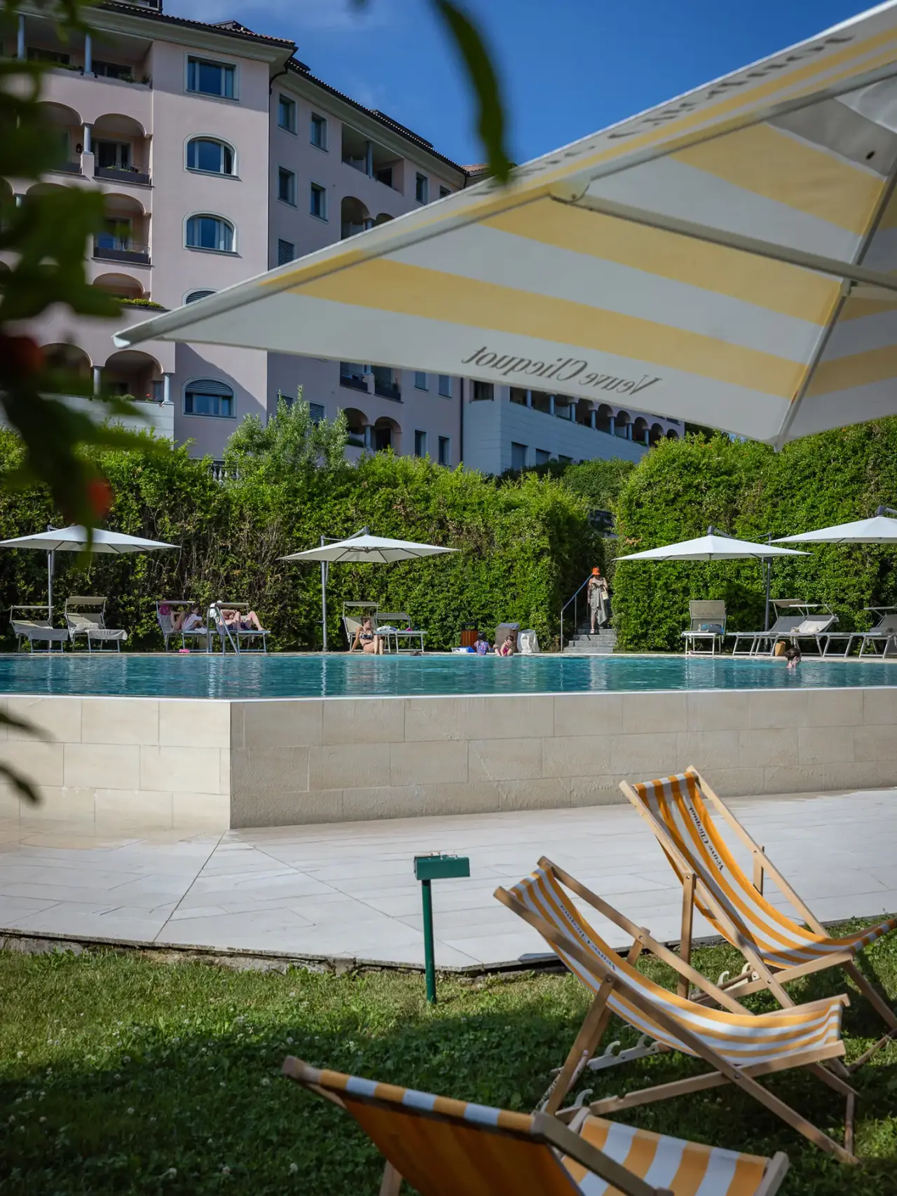 Aussenpool des Resort Collina d'Oro mit Sonnenliegen, Schirmen und Hotelgebäude im Hintergrund unter blauem Himmel.