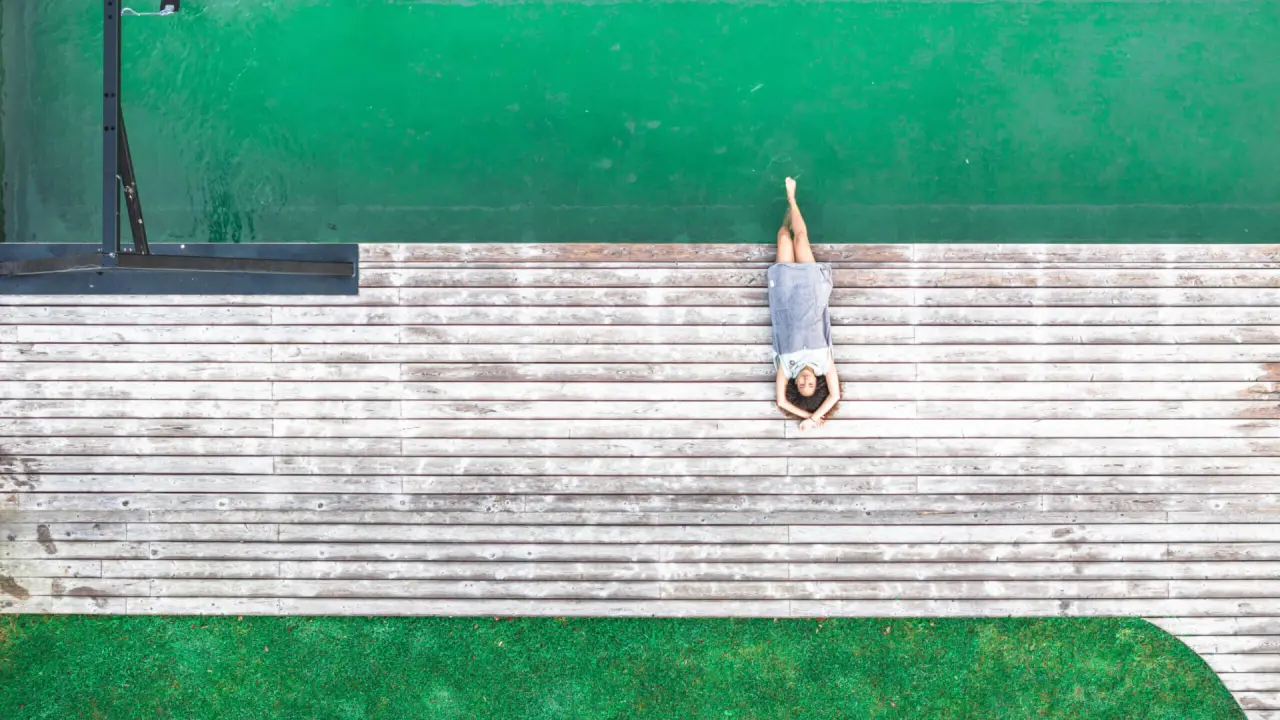 Entspannte Frau auf Holzterrasse am smaragdgrünen Naturpool des Hotel Das Falkenstein Kaprun. Idyllische Erholung im Freien.