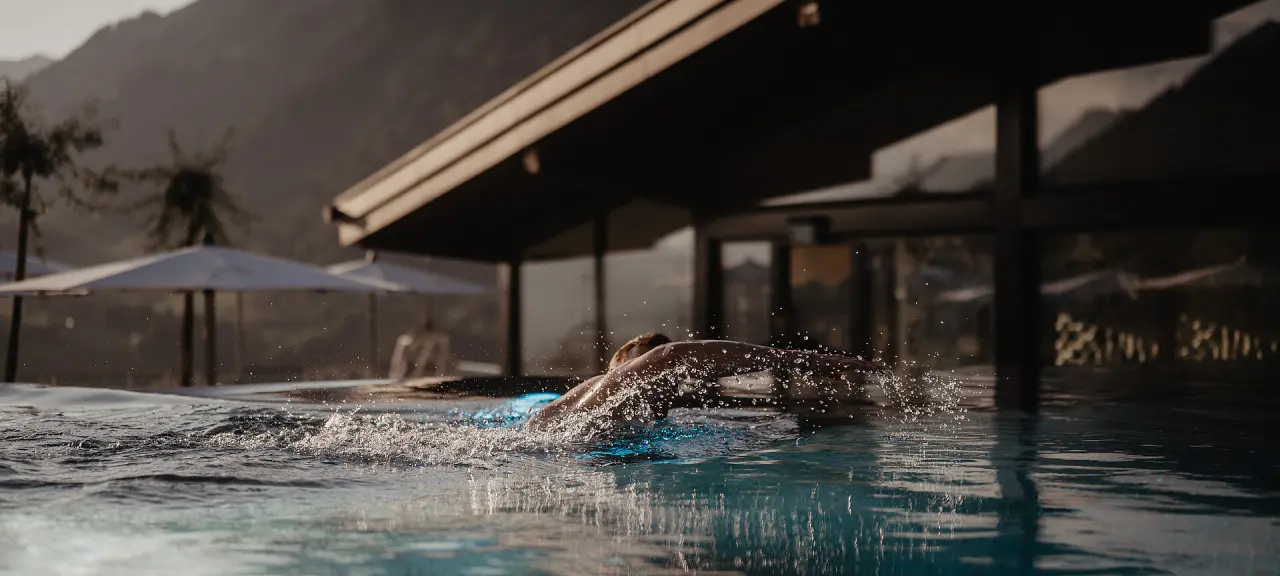 Gast schwimmt im Infinity Pool des Hotel Berghof in St. Johann, Salzburg, mit Bergblick bei Sonnenuntergang und Wasserspritzern.