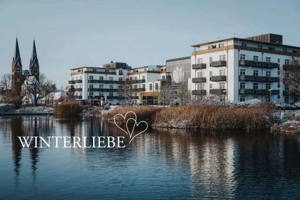 Winterliche Aussenansicht des Resort Mark Brandenburg am Ruppiner See mit verschneiter Uferlandschaft und Kirche.