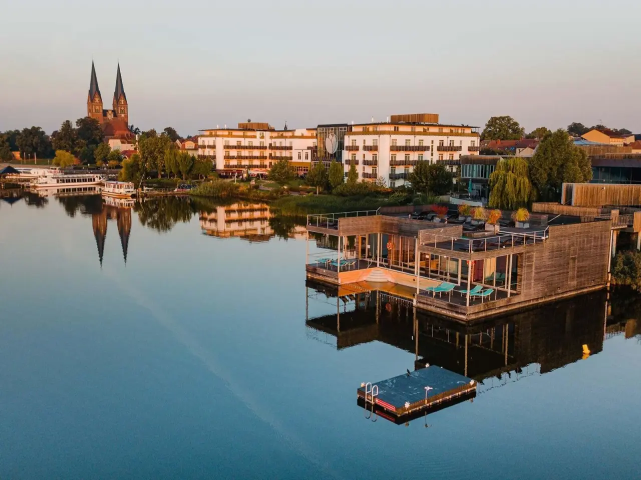 Malerische Luftaufnahme des Resort Mark Brandenburg am Ruppiner See mit schwimmenden Suiten und Hotelgebäude bei Sonnenuntergang.