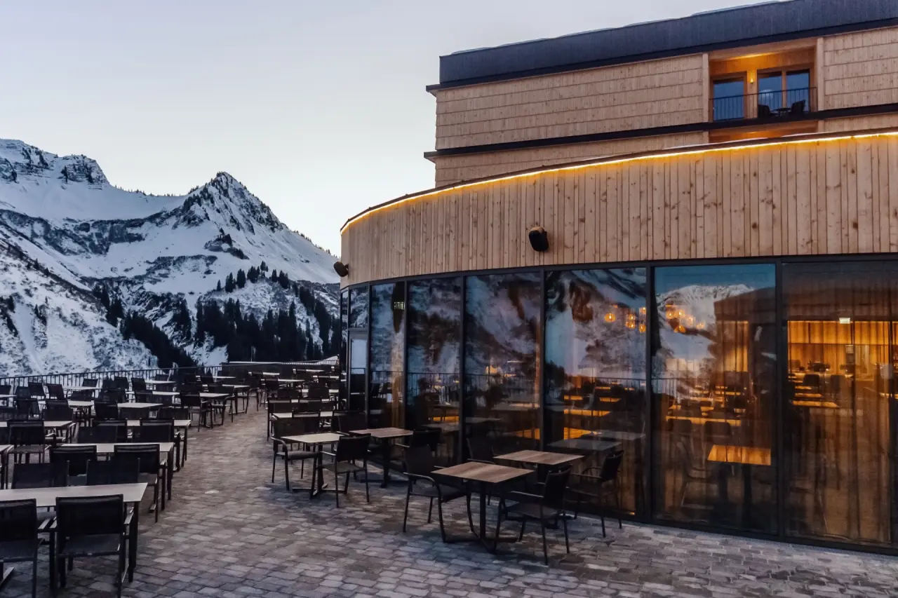 Modernes Alpenstern Panoramahotel mit beleuchteter Terrasse und verschneitem Bergpanorama in den Alpen.