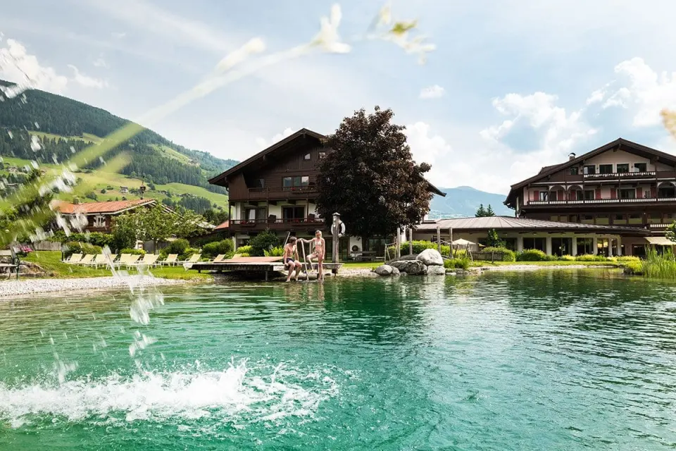 Naturbadeteich mit Holzsteg und Blick auf das Wanderhotel Kirchner und die umliegende Berglandschaft.