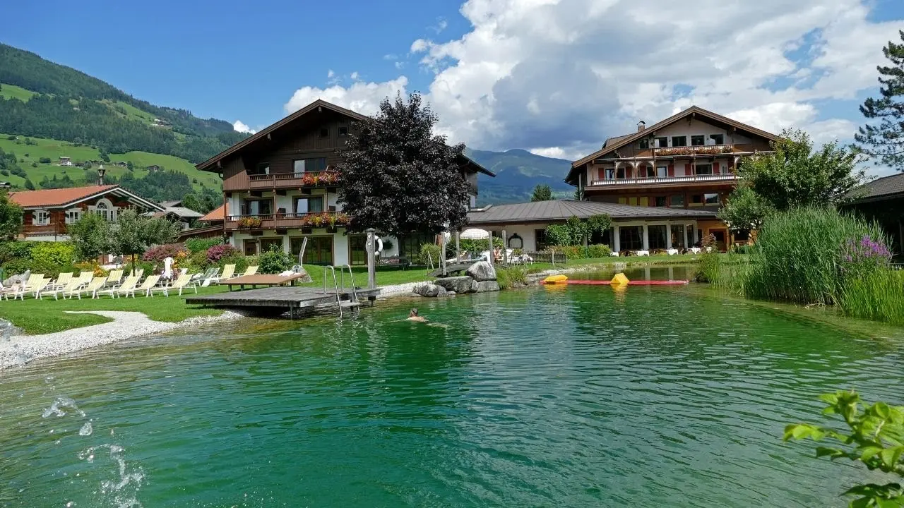 Natürlicher Schwimmteich mit Liegewiese und Alpenpanorama vor dem Wanderhotel Kirchner an einem sonnigen Tag.