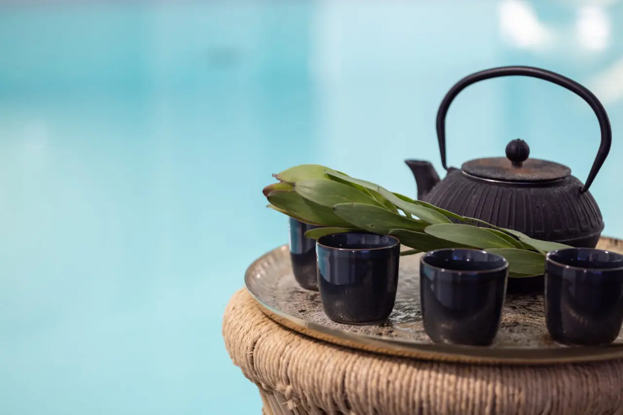 Gusseisen-Teekanne mit Tassen und grünen Blättern auf Tablett, im Hintergrund blauer Pool im Tanneck Golf & Spa.