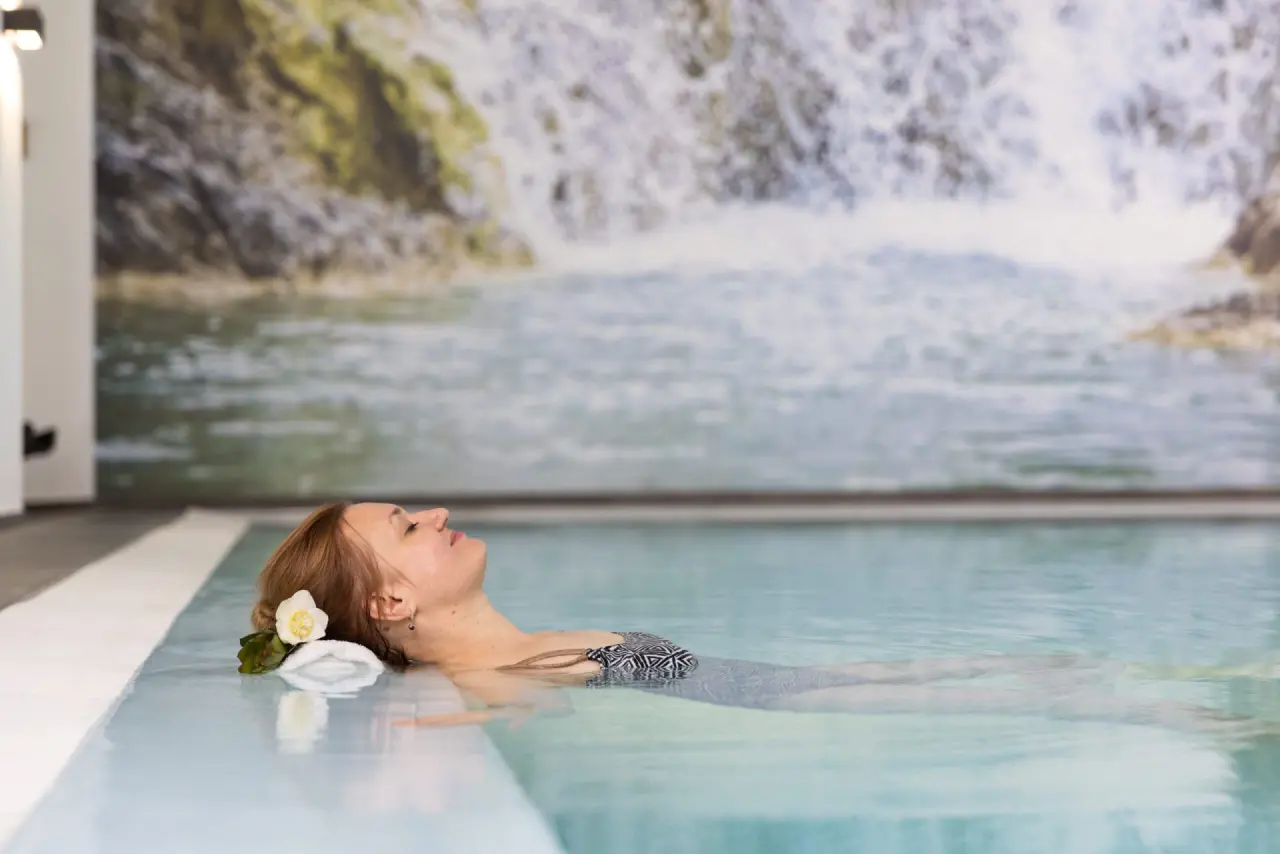 Entspannte Frau mit Blume im Haar schwebt im Pool des Hotel Tanneck Golf & Spa, umgeben von einer beruhigenden Wasserfall-Wand.