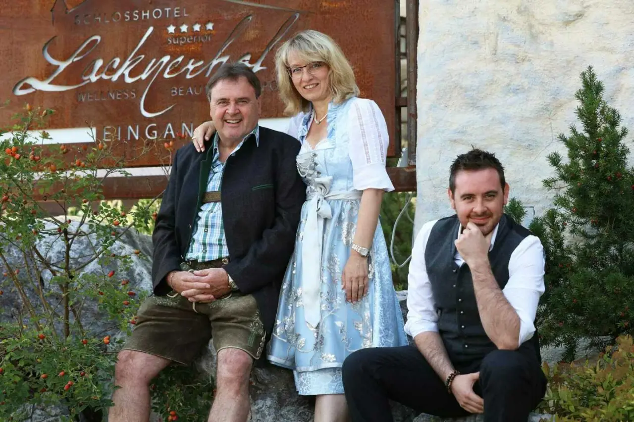 Familie Lacknerhof, die Gastgeber des Schlosshotel Lacknerhof****S, lächeln vor dem Hoteleingangsschild in Tracht.