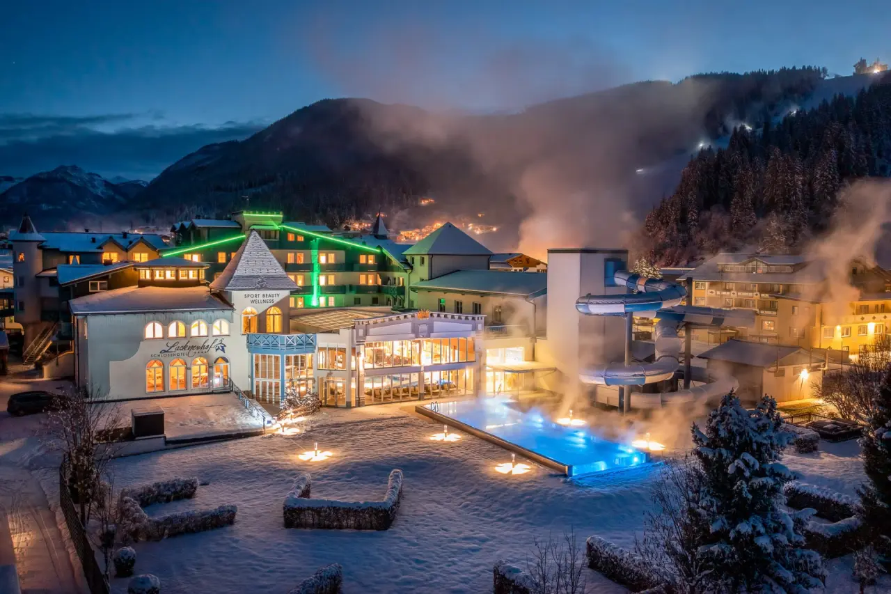 Winterliche Aussenansicht des Schlosshotel Lacknerhof****S mit beleuchtetem Aussenpool und verschneiter Berglandschaft bei Nacht.