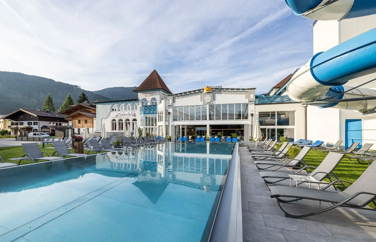 Großer Außenpool mit Liegewiese und Wasserrutsche vor dem Schlosshotel Lacknerhof****S in den Bergen.