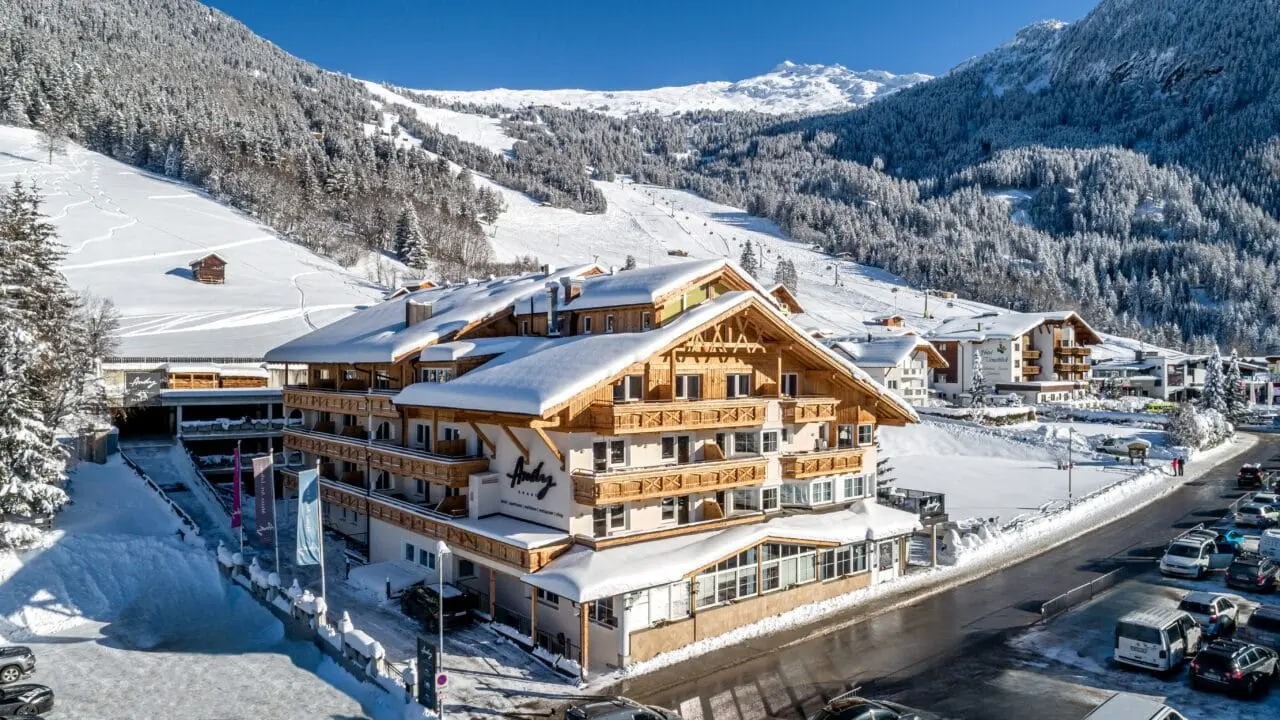 Winterliche Aussenansicht des Hotel Andy mit verschneiten Dächern und Balkonen vor einer beeindruckenden Bergkulisse.